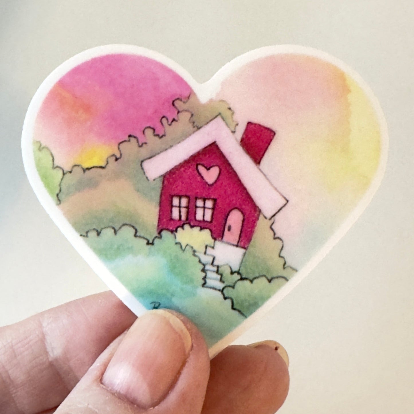 Heart-shaped sticker with a house design held by a hand against a light background