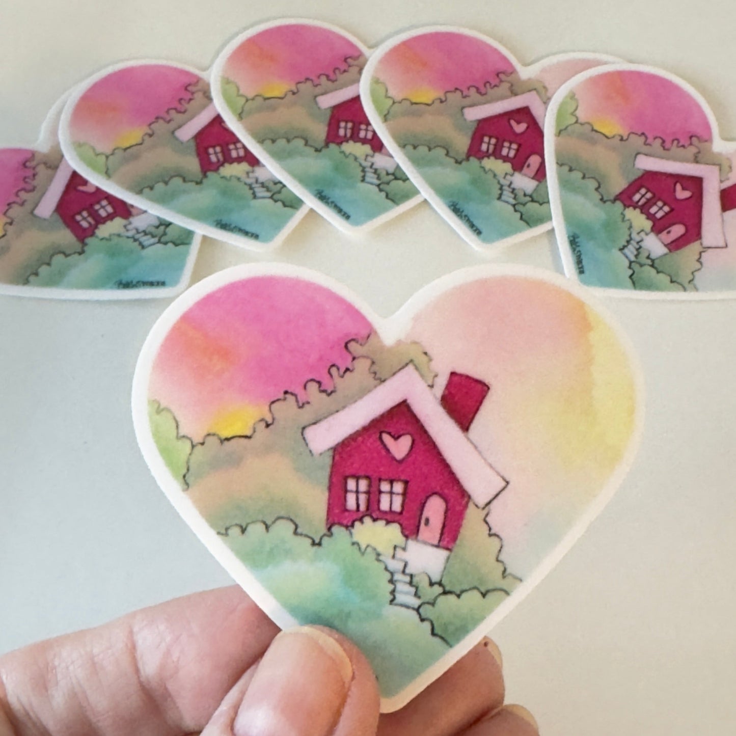 Heart-shaped sticker with a pink house design held by a hand on a light background