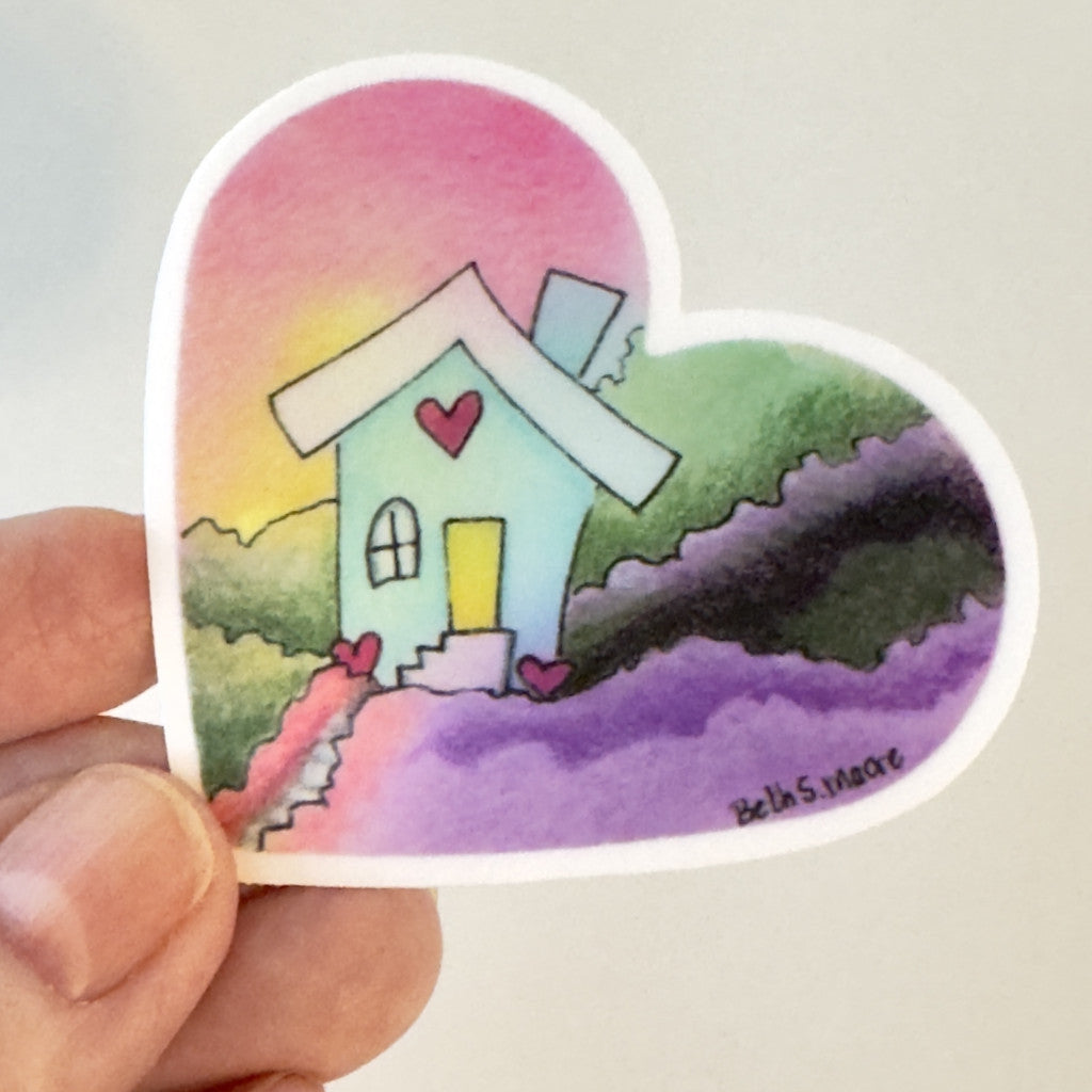 Heart-shaped sticker with a colorful house design held by a hand on a light gray background