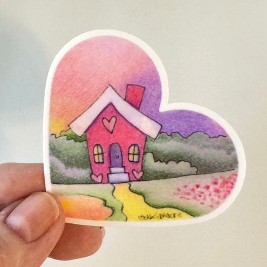 Heart-shaped sticker with a pink house design held by a hand against a light gray background