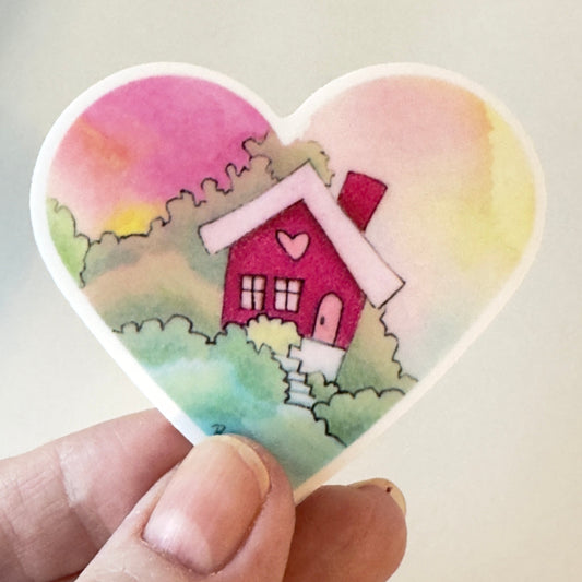 Heart-shaped sticker with a house design held by a hand against a light background