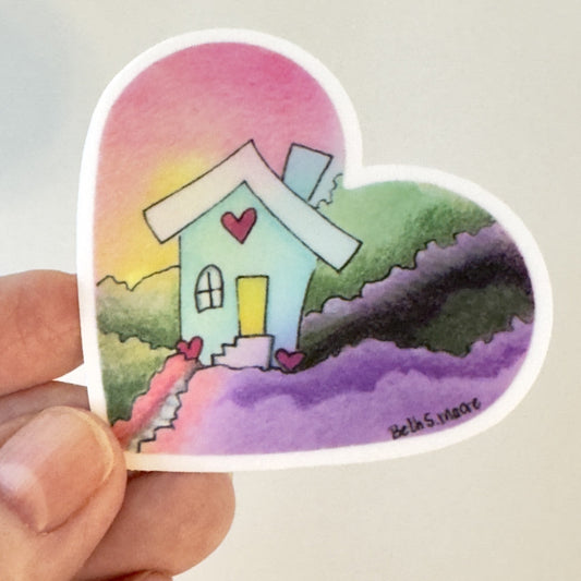 Heart-shaped sticker with a colorful house design held by a hand on a light gray background